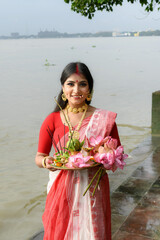 Portrait of a beautiful young Indian woman in a traditional saree, wearing gold jewellery and bangles, holding a plate filled with religious offerings in front of the river Ganges