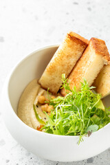 Bowl of hummus topped with chickpeas, fresh salad greens, and toasted bread slices on a light stone surface. Bright, healthy vegetarian meal presentation.