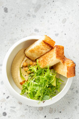 Bowl of hummus topped with chickpeas, fresh salad greens, and toasted bread slices on a light stone surface. Bright, healthy vegetarian meal presentation.