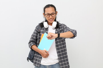 Young Asian college student wearing casual clothes, glasses, backpack, holding books, looking at watch with happy expression, punctual concept, isolated on white background.