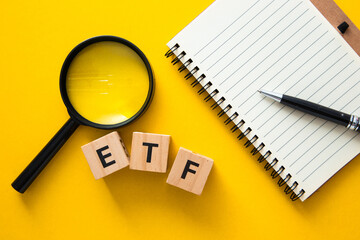 Magnifying glass is showing etf word on wooden cubes, notepad and pen on yellow background