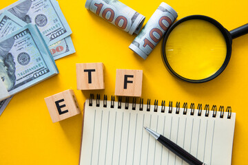 Exchange traded fund concept with etf wooden cubes, notepad, pen, magnifying glass and us dollar banknotes on yellow background