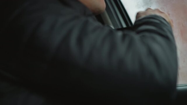 Close up of car buff wearing black winter coat pulling seat belt across torso while seated in vehicle cabin beside fogged window emphasizing safety ritual and hand gesture during cold commute