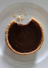 Close-up of a chocolate tart bitten into on a white plate. Its crust is golden, its filling rich with chocolate, and crumbs scattered around.