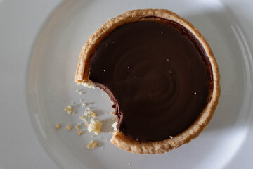 Close-up of a chocolate tart bitten into on a white plate. Its crust is golden, its filling rich with chocolate, and crumbs scattered around.