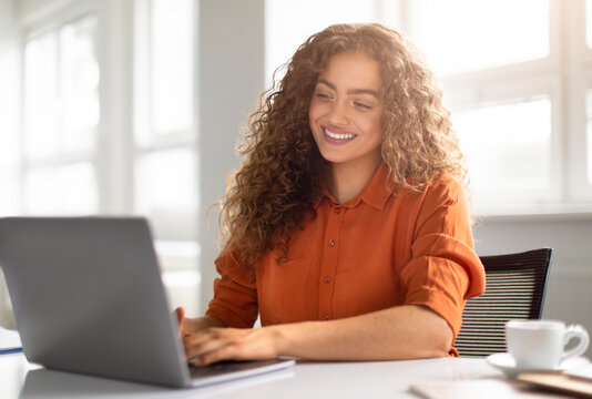 A radiant young woman in an orange shirt works on her laptop with a beaming smile in a sunlit room