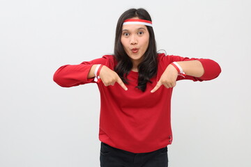 Indonesian woman celebrating the independence day of the Republic of Indonesia, surprised expression looking at the camera while pointing down to empty space, standing isolated on white background.