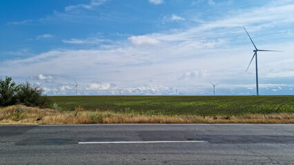 Wind turbines on green agricultural field near asphalt road under blue sky with clouds. Renewable energy landscape.