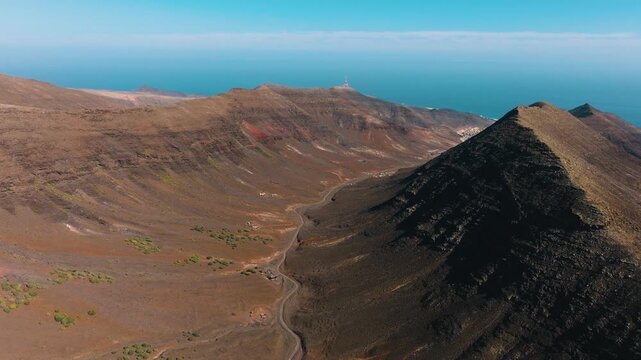 Aerial drone view of Barranco del Ciervo canyon near Solana Matorral, Fuerteventura, with mountain ridges and desert terrain, looking toward the town