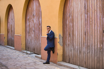 A young, handsome, dark-haired latin man wearing a suit and tie and black lace-up shoes rests his leg against a wall and smokes a cigarette. 31 May, World No Tobacco Day.