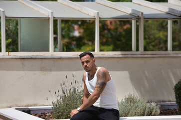 A young, handsome gay man with a beard, tattoos and earrings, wearing a white tank top and black trousers, sitting on a flower bed. The man is relaxed and resting with a serious expression.