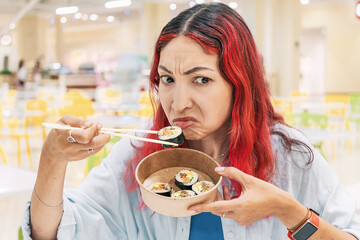 Young woman grimacing while eating kimbap or sushi with chopsticks in a bustling food court,...