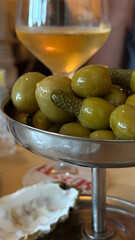Green olives and pickles served in a stylish metal dish with a glass of white wine in the background, creating an inviting dining atmosphere