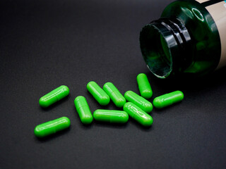 A group of bright green capsules spilled from a transparent green bottle on a black surface. The glossy two-piece capsules reflect light, creating a clean and clinical appearance