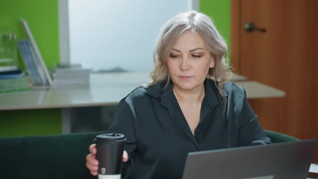 Professional woman pauses from laptop work to take sip of hot coffee before continuing typing in bright office with documents and green accent wall