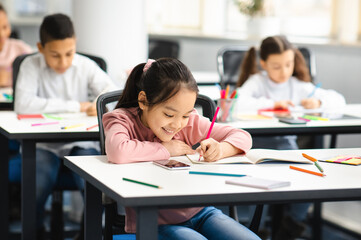 Portrait of small smiling asian girl sitting at table in classroom at primary school or kindergarten, writing or drawing in notebook. Reopening and return back to school after coronavirus quarantine