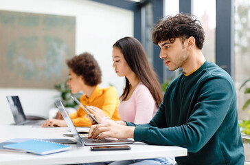 Fototapeta premium Multiracial male and female students studying for test, making homework with gadgets, focused guy using laptop, sitting at table in coworking space
