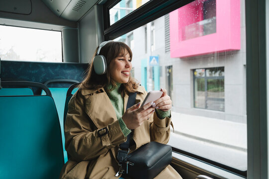 Smiling woman with wireless headphones and smart phone looking out through bus window
