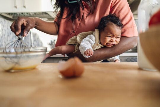 Mother whisking egg and holding crying daughter in kitchen