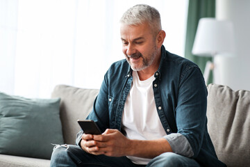 Smiling grey-haired mature man sitting on couch in living room, using mobile application on smartphone, panorama with copy space. Modern technologies in elderly people daily routine concept
