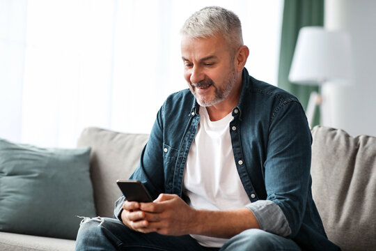 Smiling grey-haired mature man sitting on couch in living room, using mobile application on smartphone, panorama with copy space. Modern technologies in elderly people daily routine concept - Powered by Adobe