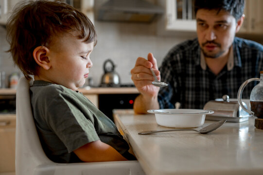 Father feeding upset son sitting at dining table in home
