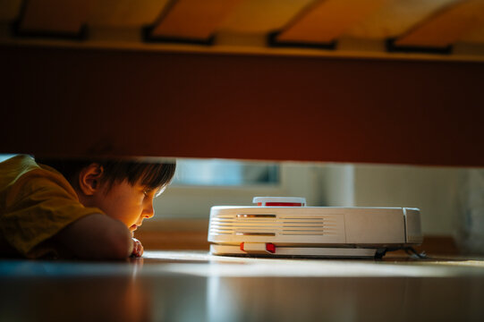 Child observing a robot vacuum cleaner under a bed in a modern home