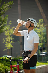 Male cyclist standing near bike and drinking water from bottle at park. Vertical