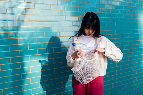 Young woman looking into mesh bag with empty plastic bottles in front of brick wall