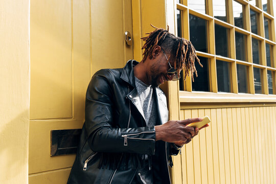 Young man with dreadlocks using smart phone by building
