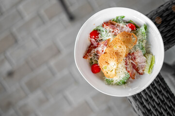 Top view of fresh Caesar salad with bacon, cherry tomatoes, croutons, and Parmesan cheese in a white bowl on wicker chair, perfect for healthy eating and food photography.