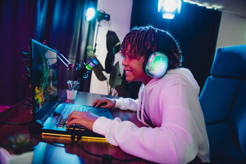 Teenager gamer using a laptop with headset in neon-lit home setup, focused on streaming or podcasting during the evening