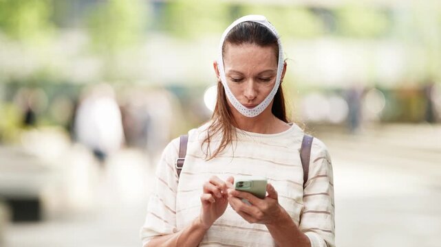 Woman in compression chin strap outdoors, focused on phone, blurred background with greenery.