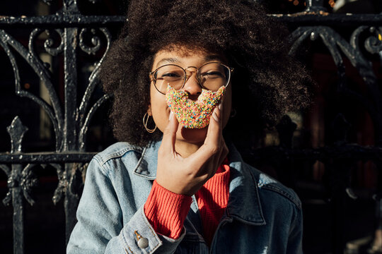 Close-up of young woman with curly hair eating donut against fence in city