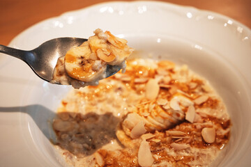 Oatmeal porridge being lifted with spoon from white bowl, topped with sliced banana and almond flakes, healthy breakfast concept