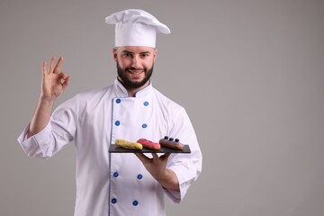 Happy confectioner in uniform holding delicious eclairs and showing ok gesture on light grey background