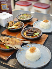 Indonesian traditional food served on a black table. Fried fish, chicken feet, spicy soup, dried beef, and rice with crispy shallots on top.