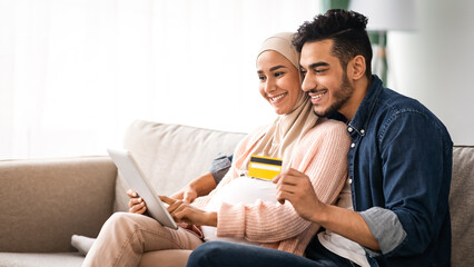 Cheerful pregnant muslim couple shopping online from home, using credit card and digital tablet. Happy expecting middle eastern family enjoying purchasing in Internet, buying baby clothes together