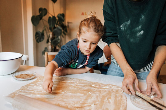 Girl sprinkling cinnamon powder on dough while helping mother in kitchen
