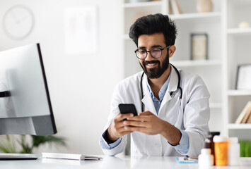 Happy young indian doctor in glasses and white coat sitting at table, using cellphone applications in clinic office. Smiling handsome general practitioner giving online mobile consultation, copy space