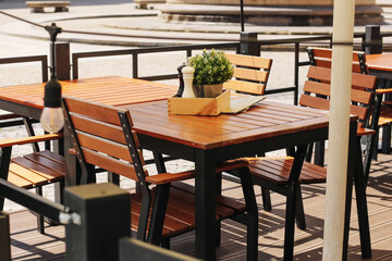 Outdoor restaurant terrace table. Wooden chairs and potted plant decor. Bright summer light and shadow. Empty patio seating arrangement. Cozy al fresco dining atmosphere. Urban cafe or bistro setup.