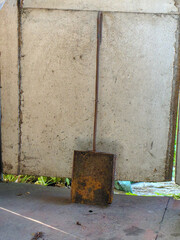 A rusty, square metal shovel leans against a textured, aged wall, outdoors.