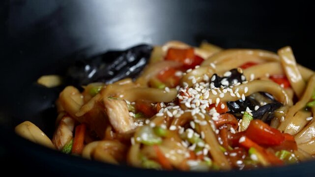 Close-up of stir-fried udon noodles with chicken, bell peppers, mushrooms, and sesame seeds in a black bowl.