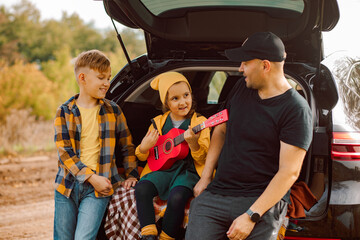 Happy dad with little cute smiling girl and boy standing near by on open car trunk. Kid resting with her family in the nature. Autumn season.