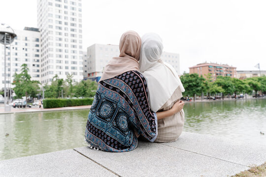 Muslim sisters sitting arm around on retaining wall by river in city