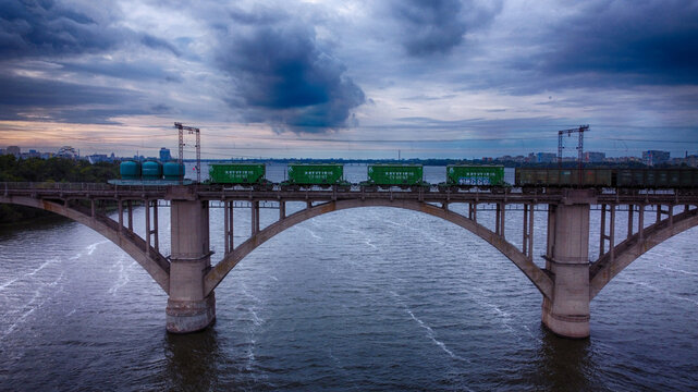 Dnipro, Ukraine - 25 September 2021: Aerial view of a train traversing the arched Amur Bridge over the tranquil waters of the Dnipro River under a dramatic, brooding sky.