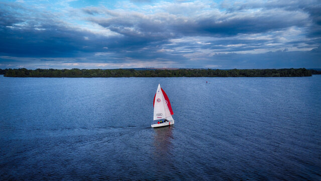 Dnipro, Ukraine - 25 September 2021: Aerial view of a lone sailboat gliding across the dark, reflective waters of the Dnipro River, its red and white sail a striking contrast against the ominous sky.
