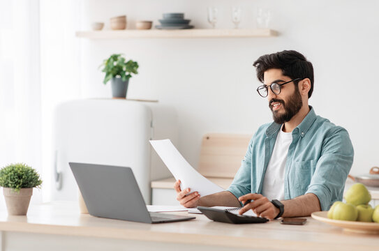 Savings and finances concept. Young arab guy using calculator and laptop computer, calculating taxes or planning budget while sitting at kitchen table at home, free space