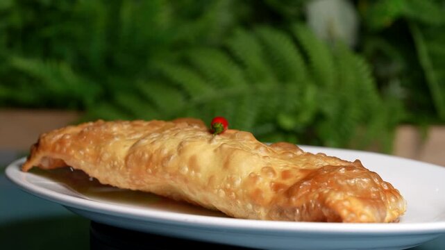 Long crispy cheburek with golden bubbles, topped with a small red chili, served on a white plate.