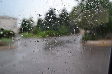 Rain drops on a window glass with blurred street and trees in the background, symbolizing melancholy, freshness, and rainy weather atmosphere